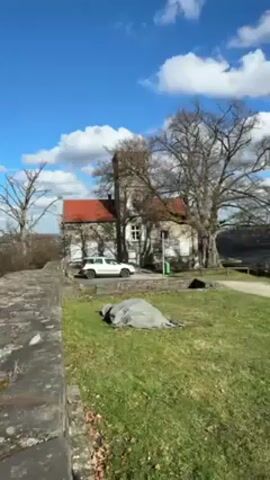 The video shows a stone building with a red roof and white window frames. There's a white car parked in front of the building, and a stone wall in the foreground. As the camera zooms in on a window, a small, cartoon-like ghost figure becomes visible inside. The ghost is white with black eyes and mouth, and it's standing on a ledge or shelf inside the window.