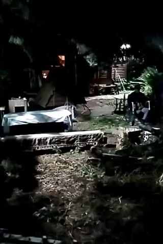 A person sits in a chair in a dimly lit outdoor area. There is a mattress and a table with a white sheet on it in the foreground. Behind the person, there are some wooden structures. The scene appears to be a backyard or a rustic outdoor space at night.