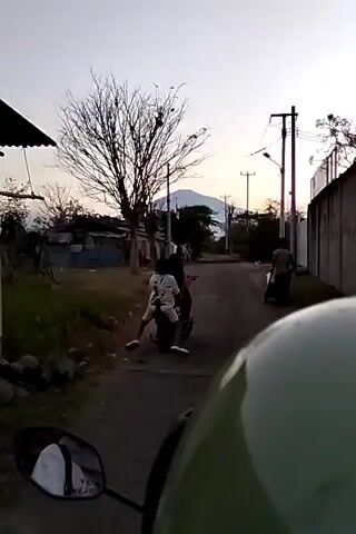 The video captures a street scene with a few people and a motorcycle. The time appears to be late afternoon or early evening, with a subdued sky. A motorcycle with two individuals is the central focus, the passenger appearing to be a young person wearing a white shirt. They are moving away from the camera. To the right, another person is visible on the road. The background includes a bare tree, some simple structures, utility poles, and a distant mountain under a pale sky. The perspective suggests the video is filmed from another moving vehicle, likely a motorcycle, given the visible mirror in the foreground. The road is unpaved, and the area seems rural or semi-urban.