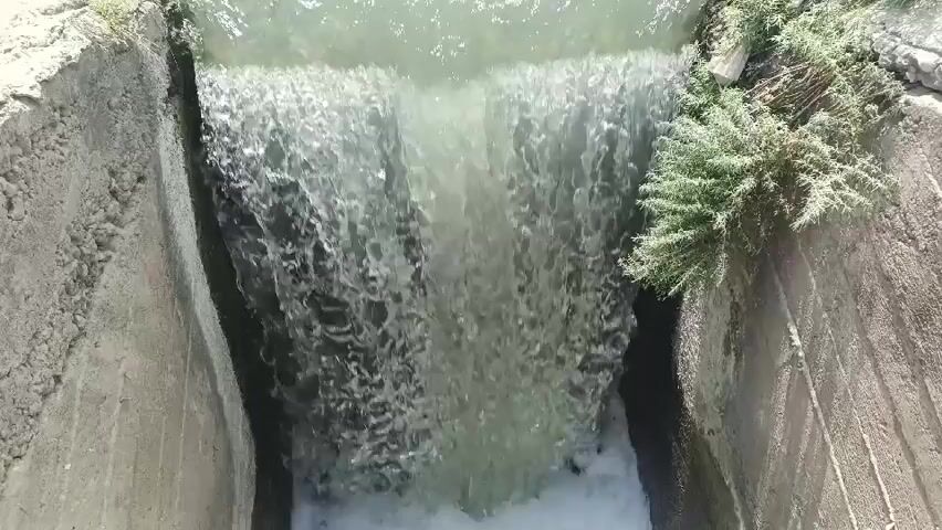 The video shows a view from above of water flowing down a concrete structure. The water appears murky and is turbulent as it cascades downwards. Green vegetation is visible on the right side of the structure, growing out of the concrete. The concrete sides of the channel are weathered and rough. The flow of water creates white foam at the bottom where it impacts. The camera angle is looking down, suggesting a drone or elevated perspective.