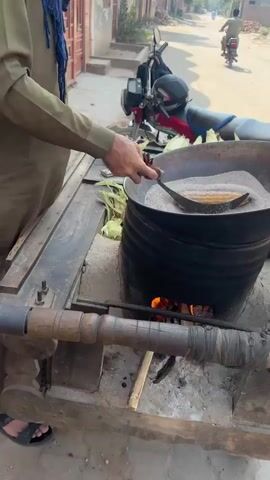A man in a brown shirt and blue scarf is cooking corn on the cob. He stands next to a cart with a metal bowl on top, filled with a dark sandy substance. A fire burns underneath the bowl. Using a flat tool, he pushes the sandy substance around the corn on the cob. The cart is parked on a street next to a red motorcycle. There are houses on both sides of the street. Another person is riding a motorcycle in the distance.