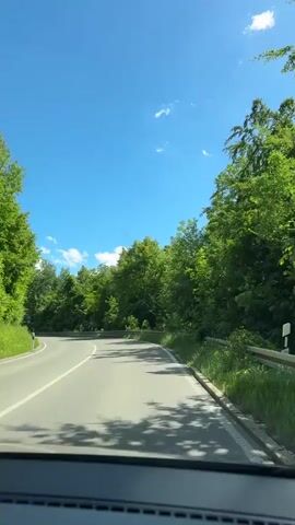 The video shows a first-person view from a vehicle driving on a paved road surrounded by lush green trees under a clear blue sky. The road curves to the left. A metal guardrail runs along the right side of the road. There are a few white clouds in the sky. There are no people visible in the immediate foreground, but two motorcycles are seen in the distance on the left side of the road, approaching the camera's vehicle. The sun is shining, casting shadows on the road from the trees.
