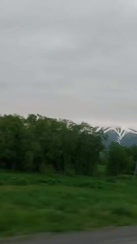 The video shows a car driving along a paved road, with a vast, green, grassy field on the right. In the distance, there are snow-capped mountains shrouded in clouds. A simple wooden cross stands prominently in the field. The sky is overcast, and the overall atmosphere is calm and serene. The camera captures the scenery from the perspective of someone inside the moving vehicle, indicating a journey through a natural landscape.