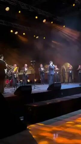 A group of men in suits are standing on a stage, illuminated by spotlights. A singer stands front and center with a microphone, singing, as other men play musical instruments like trumpets and saxophones. Smoke effects are visible in the air. Speakers are positioned on the stage, and the stage flooring has a geometric pattern. Overhead, there's visible rigging and lighting equipment. The view is from the audience, capturing the scene from a low angle.