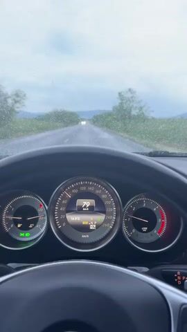 The video shows a driver's perspective from inside a car driving on a wet road. Raindrops are visible on the windshield, and the road is slick. The car is traveling at 79 km/h, as indicated on the dashboard. The car's navigation system is displayed on the infotainment screen. Headlights of an oncoming car are visible in the distance. The sky is overcast, and the surrounding landscape includes trees and fields.