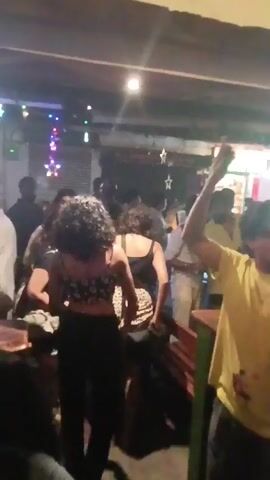 The video shows the interior of a bar or restaurant with several people. In the initial shot, people are standing, dancing, and socializing. One man in a yellow t-shirt has his arm raised in the air, appearing enthusiastic. The camera then pans to different areas, showing people seated at tables, and others standing and talking. The lighting is a mix of bright overhead lights and decorative colored lights. Christmas decorations, including stars, are visible. The overall atmosphere appears lively and festive.