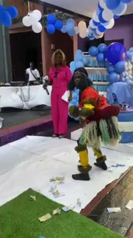 Here is a description of the video.

The video takes place at an indoor party with blue and white balloon decorations. A person in traditional African garb, including a red top, a skirt made of green and white fibers, yellow leg coverings, and a black face covering, is dancing, kicking their feet out and scattering scattered paper money across the floor. A woman in a pink jumpsuit stands nearby, smiling and holding a microphone. In the background, there's a DJ setup with a man standing behind it. A section of artificial grass is visible on the floor.