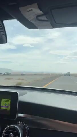 The video is shot from inside a car looking out the windshield. The car is on a highway, driving through a desert landscape. The sky is cloudy with patches of blue. In the distance, there are mountains and power lines. Other cars and a truck can be seen on the highway. A smaller vehicle with people standing in it is visible off-road. The dashboard of the car is visible in the foreground, including a screen that displays information. The overall impression is of a road trip through a somewhat desolate but scenic environment.