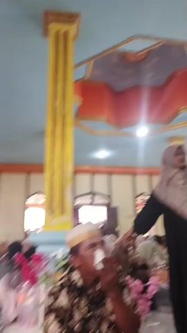 The video captures an indoor gathering, likely a ceremony or event, with a mix of individuals dressed in traditional attire. The room features ornate ceiling decorations with geometric patterns in red, orange, and blue, and tall yellow pillars. Several people are visible, including a woman in a black dress and a patterned headscarf speaking or gesturing, and men wearing peci hats and patterned shirts. In the foreground, two men are seated, one in a black and white patterned shirt and the other in a brown and black patterned shirt. In the background, other attendees are seated, some partially obscured, and there are floral arrangements, appearing to be pink flowers. The lighting suggests an indoor setting, possibly a community hall or a mosque. The camera is in motion, creating a slightly blurred and dynamic view of the scene.