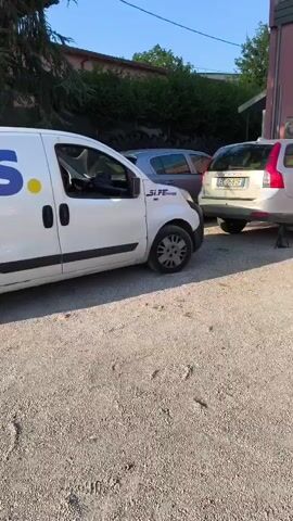 This media file captures an outdoor scene in what appears to be a workshop or garage area, possibly in Chiugiana, Italy, given the context. The time of day seems to be late afternoon or early evening, as indicated by the long shadows and clear, blue sky with some scattered clouds. The sun is shining, creating a warm, bright atmosphere.

The primary subjects are several vehicles. In the foreground, a white Audi A1 is parked, its rear facing the camera. The car has a black roof and Italian license plates reading "FE 802DJ". To the left, a white commercial van, possibly a Fiat, is also visible, with partial branding that includes "SI.FE.design". Behind the van and further into the scene, a silver Volvo station wagon is parked, with its rear jacked up, suggesting it's undergoing some form of maintenance or repair. Another car, a dark-colored hatchback, is parked behind the Volvo.

In the background, a light pink building with a large open garage door dominates the scene. Through the open door, two people can be seen inside the workshop. One person, wearing a dark t-shirt and jeans, stands near the entrance, seemingly observing or interacting with something within. Another person, appearing to be a woman in lighter clothing, is further inside the workshop. Various tools and equipment are visible within the garage space, further reinforcing the idea that this is a place of automotive work. The ground is covered in gravel. A metallic structure with a clock face is mounted on the exterior wall of the pink building. The overall mood is functional and perhaps industrious, given the presence of vehicles undergoing work.