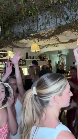 A group of young adults are dancing and celebrating in a dimly lit indoor space with lush green vines hanging from the ceiling. Several individuals are holding up their arms, some are playing a tambourine, and others are smiling and laughing. The environment appears to be a restaurant or bar with shelves of wine bottles visible in the background. The overall atmosphere is festive and lively.