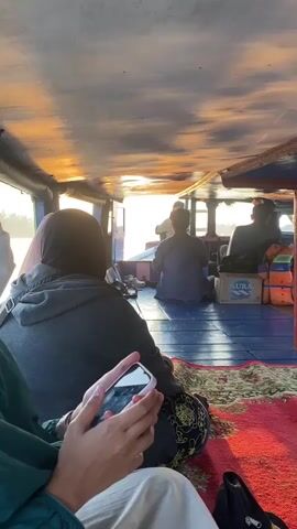 The video shows the interior of a boat moving on water. The ceiling of the boat is visible with sunlight shining through, creating orange and yellow patterns. Several people are seated inside the boat, facing away from the camera. One person in the foreground is holding a smartphone. In the background, through an opening in the boat, a calm body of water is visible, with a line of houses and trees on the distant shore. The sky is clear with the soft light of sunrise or sunset. The overall atmosphere appears peaceful and serene.