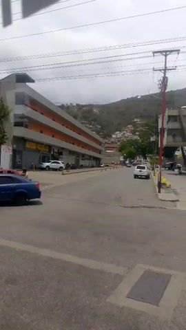 The video shows a view from a moving vehicle driving along a street in what appears to be a city or town in a hilly region. The weather is overcast. There are several cars parked along the street and other cars moving. Buildings line the street, some commercial with storefronts, and others residential further up the hill. At one point, the camera passes by a stack of shipping containers, including those with markings from Maersk and Hamburg Süd. A white police SUV passes by, and a man walks in the crosswalk. Power lines are visible overhead.