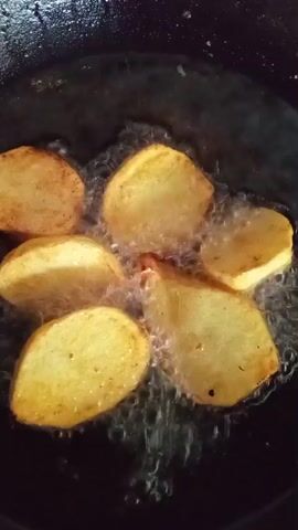 A close-up, top-down view shows potato slices being fried in a pan. The potato slices are golden brown and are bubbling in hot oil. The pan is dark and appears to be cast iron. The oil is actively sizzling and creates a lot of steam and bubbles around the potatoes.