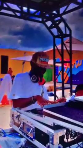 A close-up shot shows a man DJing at what appears to be an outdoor event. The DJ is wearing a white shirt, sunglasses, and a brown cap with “SPEAK” printed on it. He has headphones around his neck and is actively using the turntables. To his right is another man focused on a silver laptop. They are situated under a metal frame structure, with colorful graffiti art in the background. A few people walk around in the background, and white umbrellas are visible, suggesting an outdoor venue. The sky is partly cloudy.