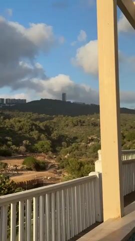 The video shows a scenic view of a hilly landscape with greenery and some buildings in the distance. The foreground features a white railing and a cream-colored pillar, suggesting the viewpoint is from a balcony or porch. The sky is partly cloudy with patches of blue. The hills are covered with trees and shrubs, and there are some dirt areas and structures visible on the slopes. In the far distance, several tall buildings and a distinct slender tower can be seen atop the hills. The light suggests it might be late afternoon or early morning due to the warm tone on the pillar.