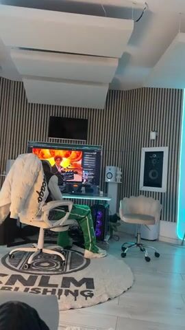 A person sits in a white office chair in front of a computer. The computer shows a video of a woman with red curly hair. The person in the chair is wearing green pants with white stripes and a white jacket. The room has striped walls and soundproofing panels on the ceiling. There is a monitor mounted on the wall above the computer monitor. Other objects in the room include a white speaker, a ceramic bunny figurine, and a white chair. The person appears to be watching the video on the computer screen.