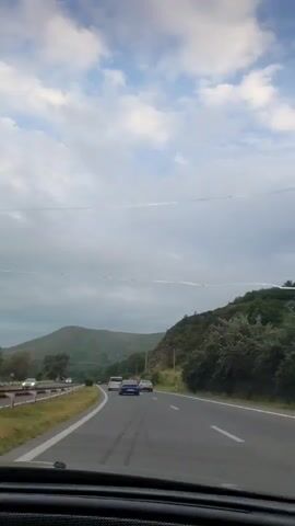 A POV shot from inside a car driving on a two-lane highway. Cars are driving in the same direction. There are green hills on the right side of the road, and more hills in the distance. The sky is cloudy with some blue patches.