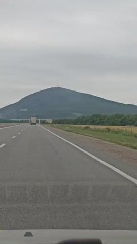 The video shows a view from inside a vehicle, likely a car, traveling on a highway. The road has at least two lanes in the direction of travel, marked with a white line. The sky is overcast. To the right of the road, there is a field of dry grass or wheat, followed by a line of trees. In the distance, a large, rounded mountain is visible, with what appears to be a structure, possibly a radio tower, at its peak. Several trucks are seen driving on the highway in the same direction. The view is slightly blurry, suggesting motion.
