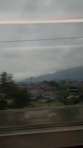The video, taken from a moving train, shows a landscape featuring residential buildings with red rooftops set against a backdrop of green fields and tree. Distant mountains are shrouded in mist under a cloudy sky. Power lines and communication towers are visible in the foreground, interspersed with the houses. The camera pans slightly as the train moves, offering different perspectives of the scenery.
