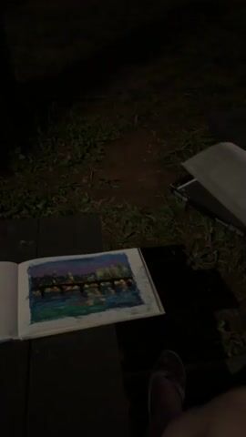 The video shows a sketchpad with a drawing of a city skyline with reflections in water. The sketchpad is on a dark surface. In the background, a grassy area is visible, with trees and lampposts. Further in the background, there is a body of water with a bridge and city lights in the distance. The scene is illuminated by artificial lights and appears to be at night. The camera is positioned low, looking slightly upwards.