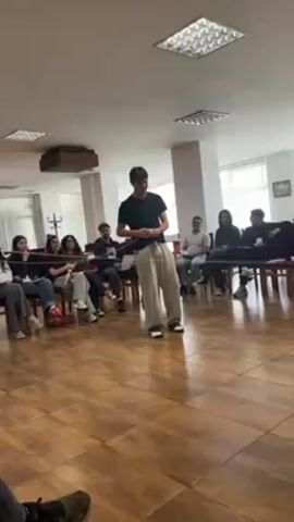 A young man stands in a circle of seated people, holding a length of string. Another person approaches and the two hold the string together, raising and lowering it. The seated people are partially encircled by the string. The room is lit by fluorescent ceiling lights. The floor is brown patterned tiles. There are approximately 15 people, mostly young adults.