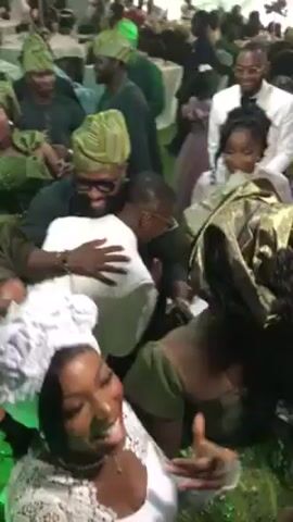 The video shows a gathering of people, likely at a wedding or celebration, mostly dressed in green and white attire. The camera is held by someone within the crowd. In the foreground, a woman in a white dress and headwrap looks at the camera and smiles. Behind her, other guests are dancing and interacting, some hugging. Many of the men are wearing traditional Yoruba hats called "fila." The atmosphere appears festive and lively, with background music playing.