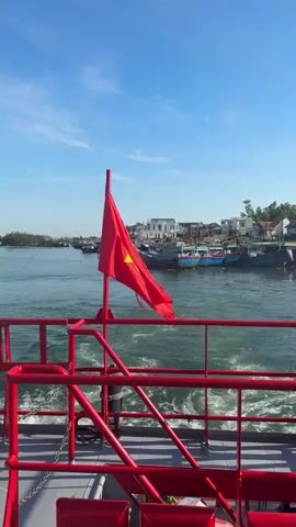 The video shows a view from the back of a red boat moving through water, leaving a wake. A red pole with a red flag is visible on the left. In the distance, a town with white buildings and green trees lines the shore. Several boats are docked or moving in the water, including some blue fishing boats and a large red passenger ferry with white lettering and stars. A white life raft is mounted on the right side of the boat in the foreground. The sky is clear and blue. The camera pans slightly to the right, revealing more of the passenger ferry and the shoreline.