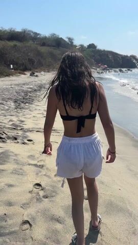 The video shows a woman walking along a sandy beach towards the ocean. She is wearing a black bikini top and white shorts. The sky is clear blue and the sun is shining. There are some rocky cliffs and sparse vegetation in the background. The water is clear with small waves lapping at the shore. A few people are visible swimming and playing in the ocean in the distance.