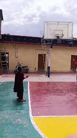 A person wearing a maroon robe stands on a colorful basketball court. They are facing a basketball hoop and are in the process of throwing a ball. The court has green, red, and yellow sections. A ladder is leaning against the basketball backboard. A building with yellow brick walls and dark trim is visible behind the court. A scooter is parked near the building. The sky is cloudy.
