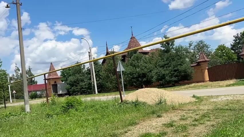 A low-angle shot captures a rural scene under a bright blue sky with scattered clouds. In the foreground, overgrown green grass and weeds dominate, with a prominent grey utility pole on the left. A bright yellow pipe runs horizontally across the frame, supported by metal poles. In the mid-ground, a road is visible, with a pile of light-colored gravel on the right side. Beyond the road, a building with distinctive red, conical roofs and wooden fencing is partially obscured by lush green trees. To the left of the main building, a smaller red-roofed house can be seen. The overall impression is one of a quiet, somewhat unkempt, but sunny day in a rural setting. There are no people or animals visible in the frame, and no actions or interactions are taking place.