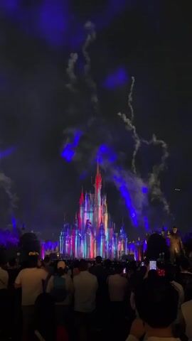 A fireworks display is taking place in front of a castle. Many people are standing in the foreground, watching the show. The castle is lit up with colorful lights and projections, and fireworks are exploding in the sky. The scene is dark, suggesting it is nighttime.