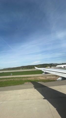 The camera is inside an airplane on the ground. The window offers a view of the runway and the plane's wing. Another airplane is also visible on the runway in the distance. Beyond the runway is a grassy area and trees. The sky is blue with scattered clouds. The airplane is stationary.