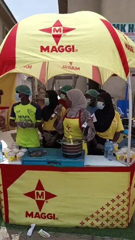 This video shows a promotional event for Maggi noodles, set up under a yellow and red branded tent with "MAGGI" logo visible on the canopy and the front of the stand. The stand is decorated with the Maggi star logo and a repeating pattern of small red stars. 

There are five individuals visible. One person in a bright yellow shirt with green accents and a hairnet and mask is standing to the left, holding what appears to be Maggi noodles. Four other individuals are standing behind the counter. Three are wearing yellow shirts, hairnets, and masks, and one is wearing a light grey hijab, a yellow apron, and a mask. The person in the hijab is holding a microphone and appears to be speaking, possibly explaining or demonstrating something. There is a portable gas stove with a pot on it on the counter, suggesting that food is being prepared. Several small yellow cups with Maggi branding are arranged on the counter. Bottles of water are also visible.

The scene appears to be outdoors, possibly a street or market, with a building in the background. The lighting suggests it is daytime, with bright sunlight. The overall atmosphere seems to be active and engaging, as indicated by the person speaking into the microphone. The people are wearing masks, which might indicate a public health awareness measure or a response to current conditions. The activity suggests a food tasting or demonstration event to promote Maggi products.
