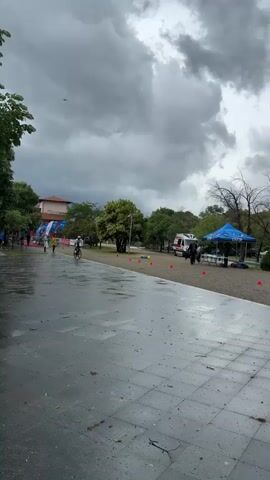 The video shows a cyclist competing in what appears to be a triathlon or cycling event on a wet, gray day. The cyclist, wearing a helmet and athletic gear, rides on a paved path marked by orange cones. In the background, there's an ambulance, a blue tent with people gathered around it, and an inflatable archway. Trees and a building are also visible. A drone is flying overhead, presumably to capture the event. The scene suggests an organized outdoor competition.