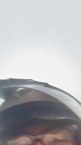 This is a first-person perspective video of a person wearing a white helmet and glasses while riding on a motorcycle. The person is looking directly at the camera. The background is blurred, indicating motion, and shows a road, grass, buildings, and power lines. Another person is visible behind the rider, also wearing a helmet and mask. The video appears to be recorded during the day, under an overcast sky.