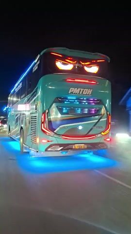 At night, a light-teal-colored double-decker PMTOH bus is viewed from the rear, driving along a dark, smooth street. The bus features distinctive red, menacing-looking decorative lights above the standard taillights, and bright blue lights are illuminated underneath the vehicle. Its license plate reads "BL 7328 AA." Other vehicles are visible in the distance ahead. The surrounding environment is dimly lit, with sparse streetlights and glimpses of structures and trees.