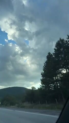 The video captures a view from inside a vehicle moving along a road. The sky is filled with dramatic, dark gray clouds, with occasional breaks revealing patches of blue sky and sunlight. The landscape outside includes rolling hills covered in dense green trees and foliage. A road barrier is visible along the side of the road. There are also a few traffic signs, including a triangular warning sign with a circular symbol and a speed limit sign indicating 100. The overall impression is of a slightly overcast or stormy day, with the vehicle traveling through a natural, somewhat rugged environment.