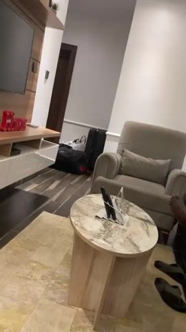 The video begins with a static wide shot of a living room. The room contains a television mounted on a wooden panel with a small plant on top, a low media console, a gray armchair, and a marble-topped round side table. There are suitcases and bags clustered near a doorway. The camera then pans downward, focusing on the floor. The floor is dark brown with a wood-like tile pattern. A pair of yellow flip-flops with black straps and a cartoon design are visible near the bottom of the frame. Part of a beige rug is also visible.