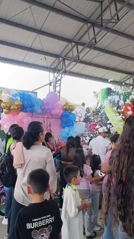 The scene is a crowded outdoor birthday celebration taking place under a metal roof. A large inflatable slide is set up, and a foam machine is actively spraying foam into the air, creating a playful atmosphere. A colorful balloon arch adds to the festive ambiance. Many children and adults are present, some are watching the foam, while others are interacting with each other. The background reveals buildings and foliage, suggesting an urban or suburban location.