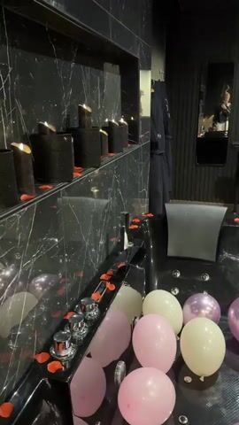 The video pans across a dimly lit, decorated room. In the foreground, a large black bathtub is filled with pink and white balloons and scattered red rose petals. To the right, the room opens up to reveal a bed covered in rose petals, with several candles lining the floor around it. Further to the right is a red massage table adorned with rose petals. The background shows black curtains, a tall black shelf unit, and on the far wall, a black X-shaped structure with restraints. A black chaise lounge chair is also visible. The overall impression is of a romantic or intimate setting.