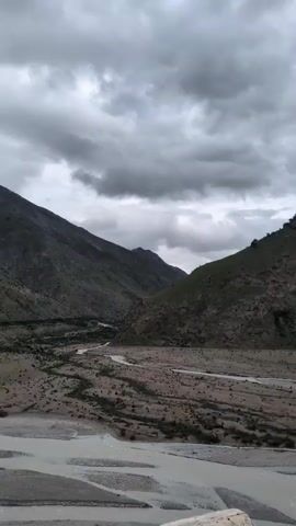 The media file depicts a vast mountain valley with a winding river flowing through it. The scene is characterized by rugged, barren slopes on either side, with sparse vegetation dotting the landscape. The sky is overcast with dramatic, heavy grey clouds, suggesting a moody or potentially stormy weather condition. The light is diffused, indicating it might be late afternoon or a heavily clouded day.

The foreground shows a wide, shallow river with multiple channels and gravelly banks. The water appears milky or muddy, possibly due to recent rainfall or erosion. Further into the valley, the river narrows and snakes through the terrain. The mountains are steep and imposing, with some appearing to have patches of snow or lighter rock near their peaks. The overall impression is one of raw, untamed nature and a sense of isolation.

There are no visible people or man-made objects in the main part of the frame, other than a small section of what appears to be a concrete structure with some graffiti at the very bottom right of the image. The lack of clear human presence reinforces the feeling of wilderness. The time of day is indeterminate due to the overcast conditions, but the shadows, if any, are soft. The location is identified as Darcha, India, which is known for its high-altitude Himalayan landscapes, consistent with the visual elements presented. The mood conveyed is solemn, expansive, and somewhat dramatic due to the weather and the sheer scale of the mountains.