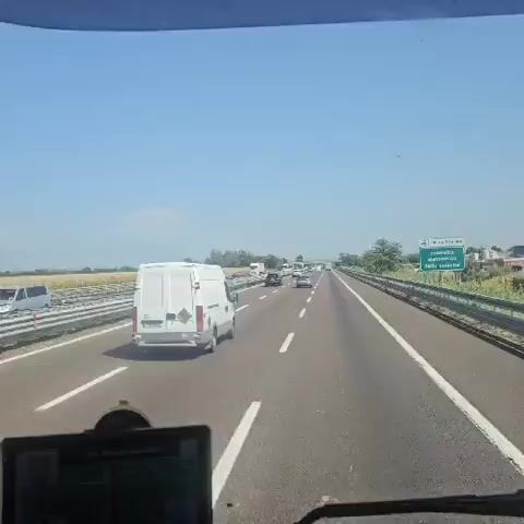 The video shows a view from a vehicle traveling on a multi-lane highway on a sunny day. Traffic is moderate, with several cars, vans, and trucks visible. The highway has concrete barriers on the sides, and agricultural fields are visible beyond the barriers to the left. A digital device, possibly a GPS or dashcam, is mounted on the dashboard in the foreground. The sky is clear and blue. There are no people visible outside of the vehicles. The scene appears to be in a rural or semi-rural area.