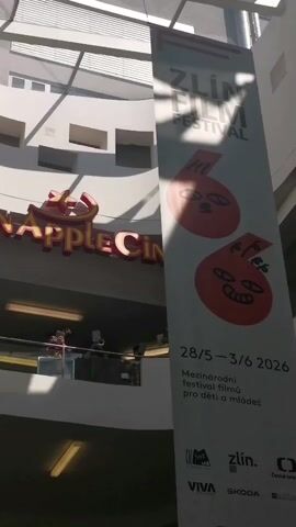 The video shows a shopping mall interior. The camera is angled upward, revealing the "Golden Apple Cinema" sign. Below the cinema, on an upper level, there are several people playing instruments, likely brass. To the right of the frame, a large banner advertises the Zlin Film Festival, which is stated to occur from May 28 to June 3, 2026. The banner contains abstract cartoon-like drawings of faces in red circles. The mall appears to be well-lit, with natural light coming in from the top of the building.