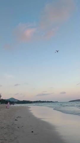 The video shows a beach scene at dusk. The sky is a light blue with pinkish clouds and an airplane is flying overhead. The beach has light-colored sand and gentle waves are lapping at the shore. People are wading in the water and walking along the beach. There is a small island visible in the distance.