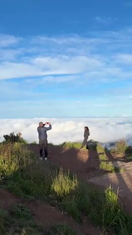 A man is taking a photo of a woman on a hill overlooking a sea of clouds under a partially cloudy sky. The man is wearing dark pants, a light blue jacket, and a baseball cap. He is holding up his cell phone to take the picture. The woman is wearing dark pants, a black jacket, and sunglasses. She is standing on the edge of the hill, posing for the photo. The hill is covered in grass and some small bushes. The sky is blue with white clouds, and the sun is shining.