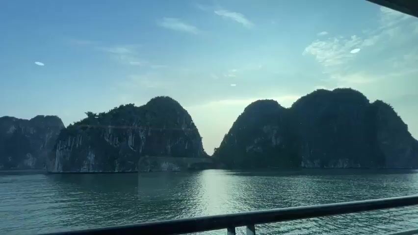 The video shows a wide shot of two large, lush green islands with steep, rocky sides rising out of calm, green water. The sky above is a clear, pale blue with a few wispy clouds. The viewpoint appears to be from the deck of a boat, with a dark railing visible in the foreground. The sun is not directly visible, but the lighting suggests it is either early morning or late afternoon, casting a soft glow on the scene. There are no people visible.