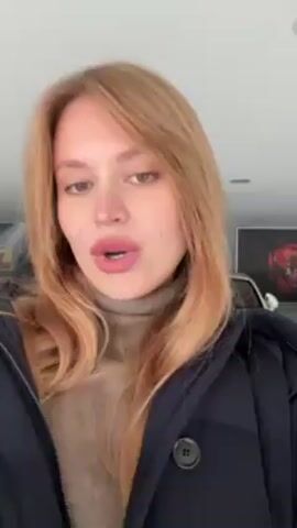 A woman with blonde hair, wearing a beige turtleneck and a black jacket, stands in a well-lit garage and says, "Hey guys, welcome to my garage." Behind her are luxury cars, including a yellow sports car and a white Bentley. The walls of the garage are decorated with automotive-themed artwork. She smiles and looks at the camera throughout the short video.