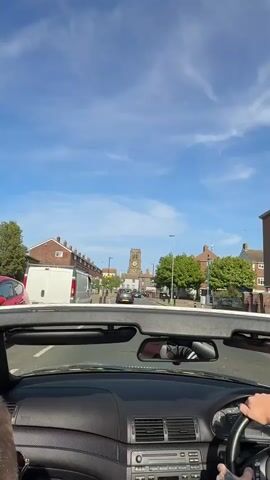A first-person perspective shows a white convertible car driving down a street in a British town. The driver's hand is visible on the steering wheel. On either side of the street, there are rows of brick houses. In the distance is a stone clock tower. The sky is blue with thin white clouds. There are other cars on the road. Music is playing.