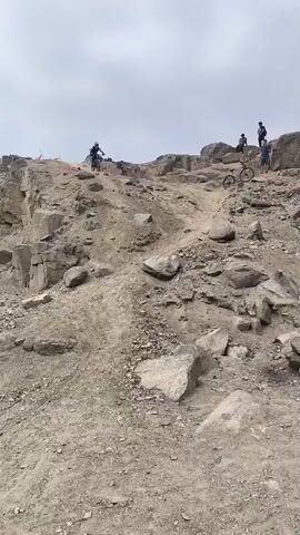 The video captures a mountain biker descending a steep, rocky, and dusty trail in Trujillo, Peru. The rider, wearing a helmet, a blue, pink, and black long-sleeved jersey, a protective vest, and knee pads, navigates the challenging terrain on a "Marin" brand mountain bike.

The landscape is characterized by an arid, barren environment with numerous large rocks and loose dirt, typical of a downhill mountain biking course. In the background, on a higher rocky outcrop, several other individuals are visible, likely observers or fellow riders.

The weather appears to be overcast, with a bright but cloudy sky, indicating daytime, possibly late morning or early afternoon. The rider demonstrates focus and skill in maneuvering through the rough and technical section of the trail, kicking up dust as they move. The footage follows the rider as they come down the slope towards and past the camera, highlighting the rugged nature of the descent. The scene conveys an intense moment of action within an adventurous sport.